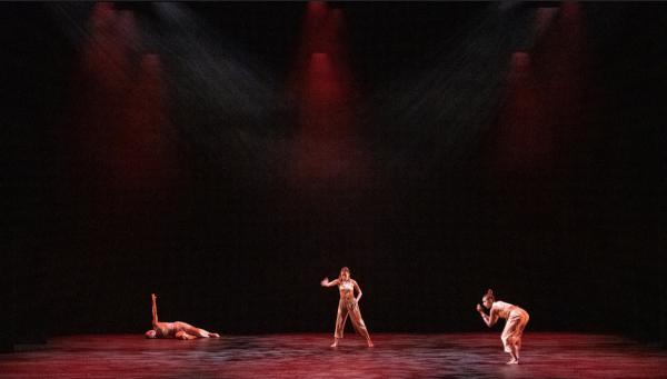 Three dancers are spread out in a black theatre stage, under red lights.