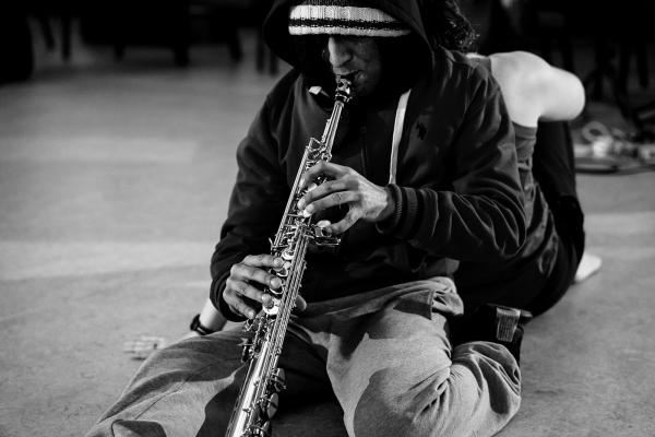 A man plays a saxophone while sitting leaning against another person's back.