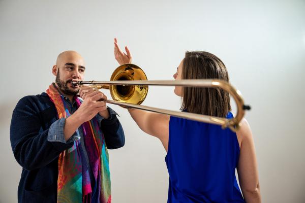 A man plays a trombone and a woman reaches a hand around the isntrument