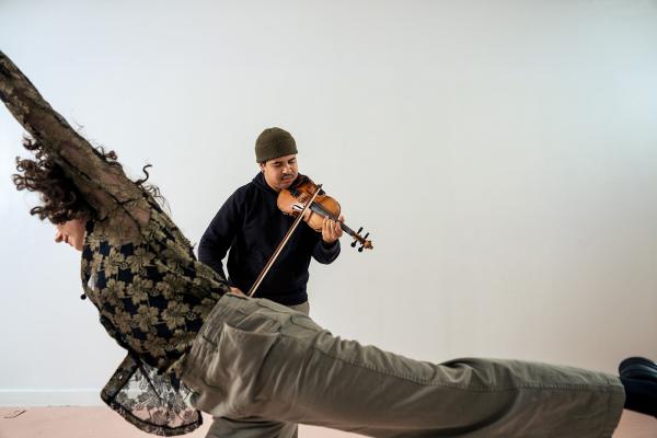 A woman reaches an arm and leg horizontally, while a man plays violin behind her.
