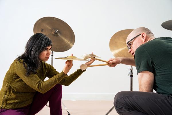 A woman kneels holidng up a cymbal as a man holds out a drumstick