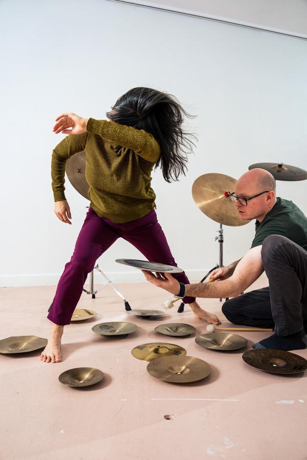 A woman dances as a man holds a cymbal, sitting among an array of cymbals