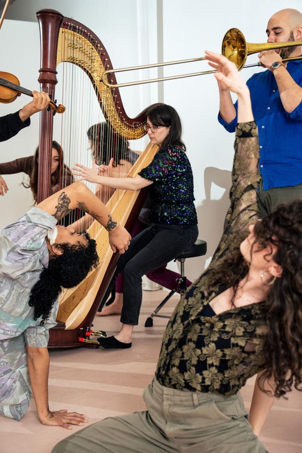A woman plays a harp, surrounded by dancers and other musicians