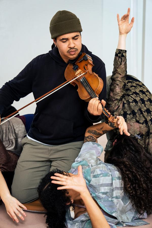 A violinist kneels, surrounded by dancers.