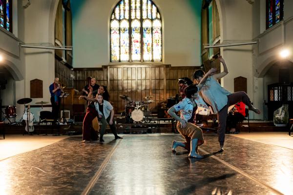 Four women dance in two duets in front of a band, in a church space.