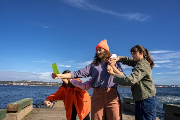 Three people dance on a wooden pier next to a bay.