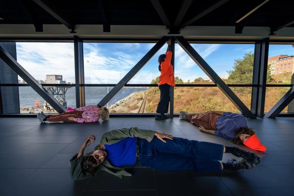 Four people take positions lying down or standing in an industrial space.
