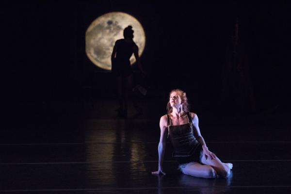 A dancer sits on stage with another dancer standing behind her in silhouette.