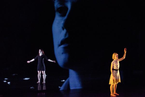 Two dancers stand with arms out in front of a photo backdrop of a woman's face.