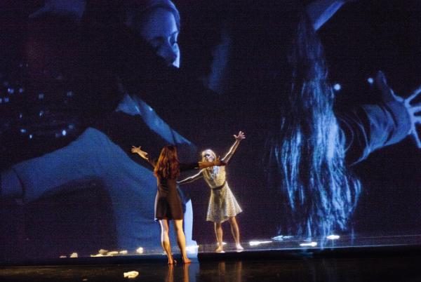 Two dancers reaching towards each other in font of a large photo backdrop.