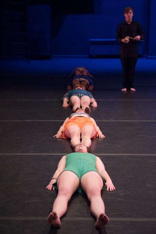 Four dancers lay lined up from head to foot, while a musician looks.