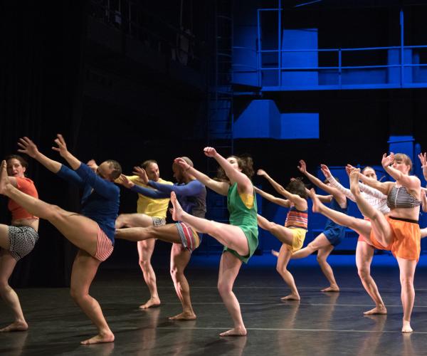 A large group of dancers in colourful clothing kick their legs high