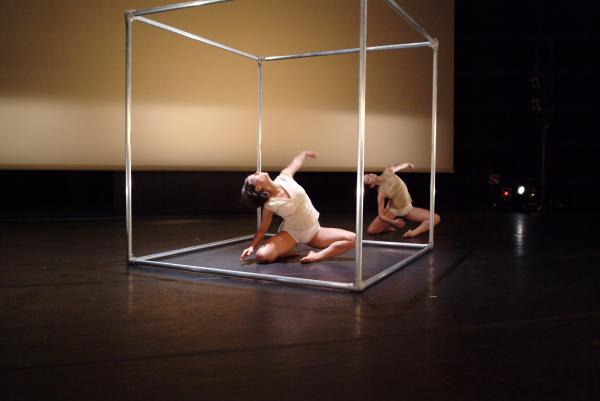 Two dancers move in the same position on the floor, one is inside a metal cube. 