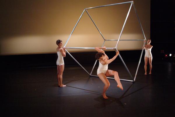 Three dancers hold a metal cube structure. 