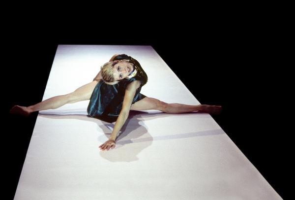 A dancer sitting on a white panel on the floor leans back to face the audience.