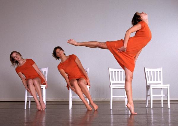 Dancers in orange dresses sit in white chairs and another dancer moves in front of them. 