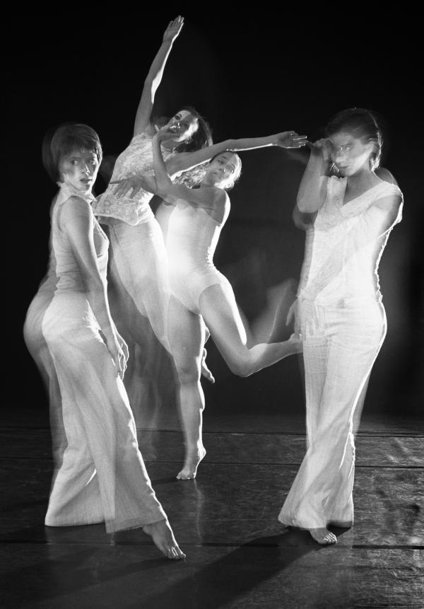 Four dancers in white costumes move in front of a black back drop. 