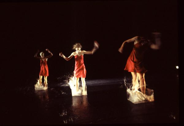 Three dancers stand in clear buckets filled with splashing water. 