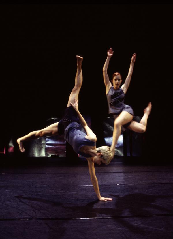 A dancer balances on one arm as another jumps in the air. 