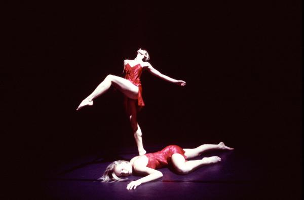 A dancer stands on one foot behind another dancer laying on the floor. 