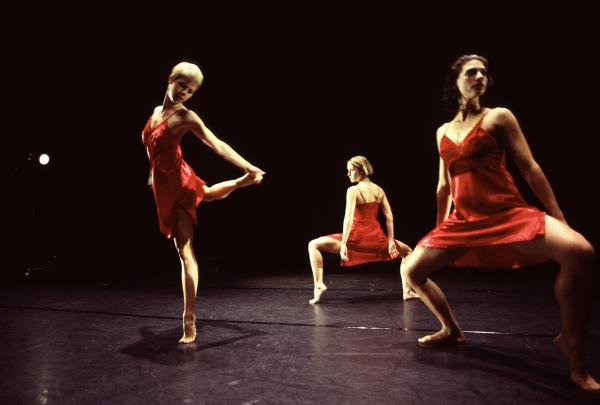 Three dancers in red dresses take different shapes in front of a black backdrop. 