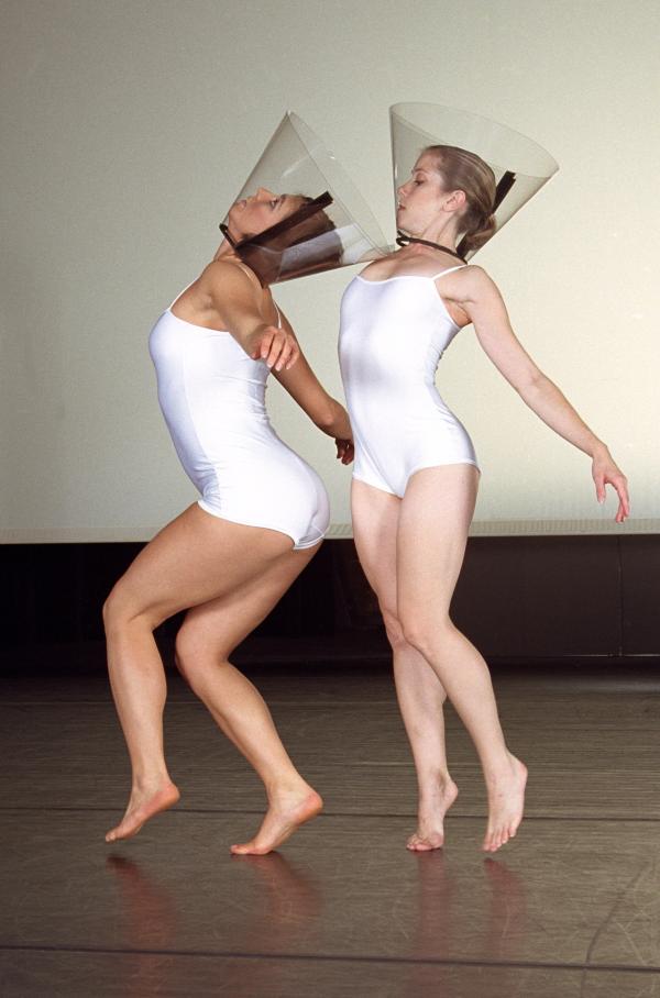 Two dancers wearing white body suits and cones around their heads face the side. 