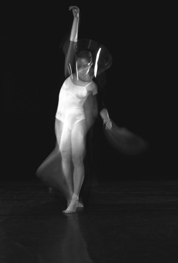 A dancer in a white body suit and transparent cone around her head stretches her arm. 