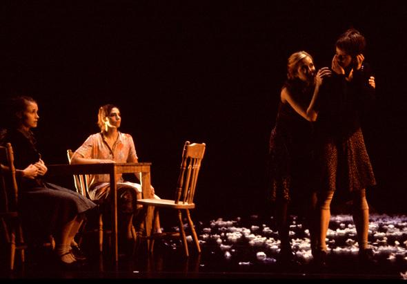 Two dancers sit at a wooden table and two dancers stand beside it. 