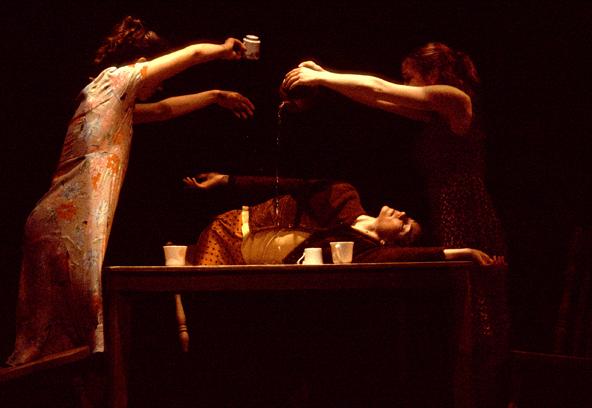 Two dancers pour tea on a dancer laying on a wooden table. 