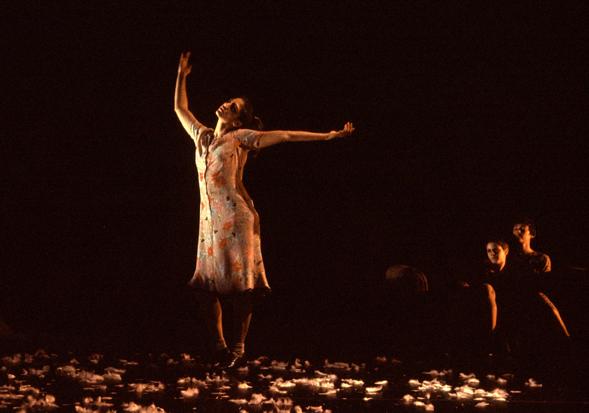 A dancer in a dress stands with arms up in front of a black backdrop. 