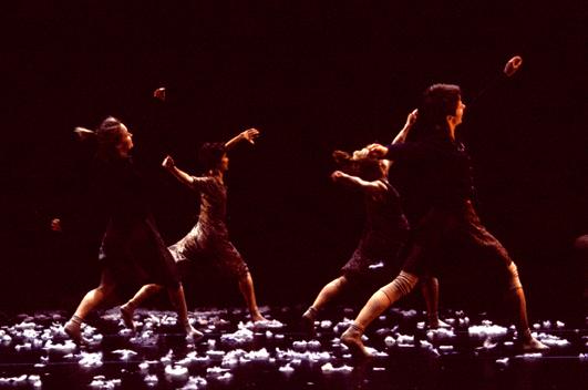 Four dancers reach in unison toward the back corner of the stage. 