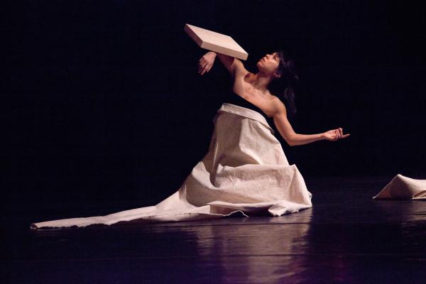 A woman kneels wrapped in canvas, balancing a block on her arm