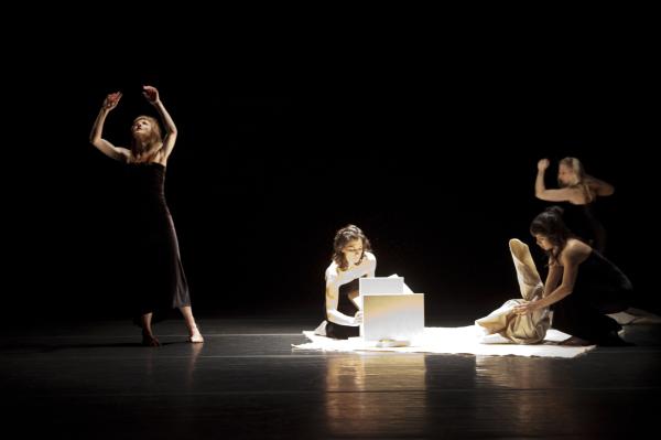 Two dancers stand in a dark theatre, and two more sit manipulating objects.