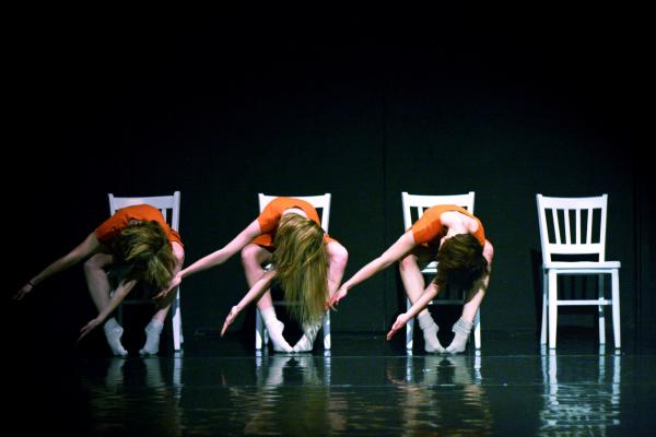 Three dancers pose leaning forward on white chairs.