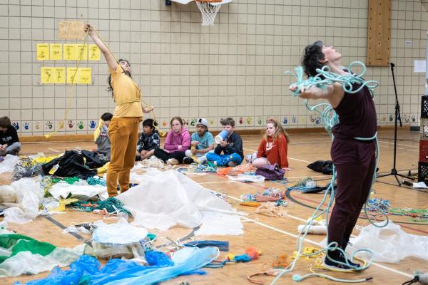 Two people stretch their arms wide, pulling at rope tangled around their chests.