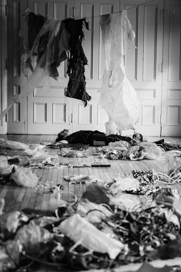 A woman lays on the floor amidst plastic debris, while more plastic dangles from a clothesline above her.