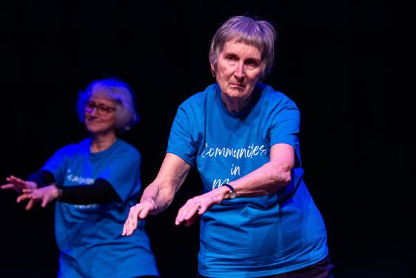 Elderly dancer in blue shirt against black background, arms reaching