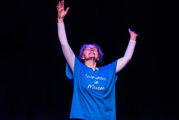 Elderly dancer in blue shirt against black background, arms reaching