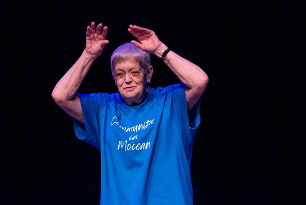 Elderly dancer in blue shirt against black background, arms reaching