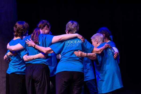 Group of elderly dancers in blue shirts embrace in a circle against black background