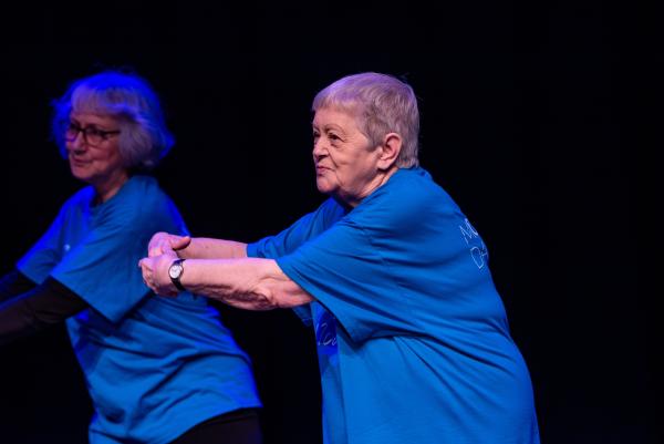 Elderly dancer in blue shirt against black background, arms reaching