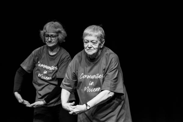 two older adults with arms in low embrace look downward on a dark stage