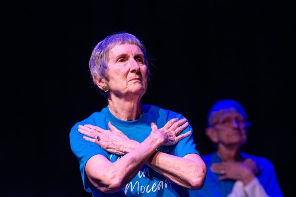 Elderly dancer in blue shirt against black background, arms crossed across chest