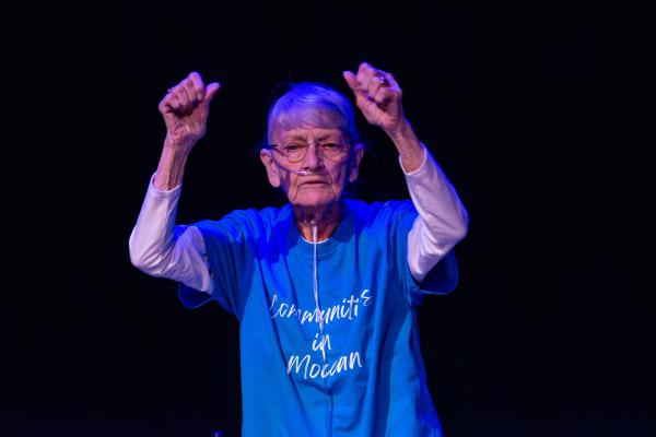 Elderly dancer in blue shirt against black background, arms reaching