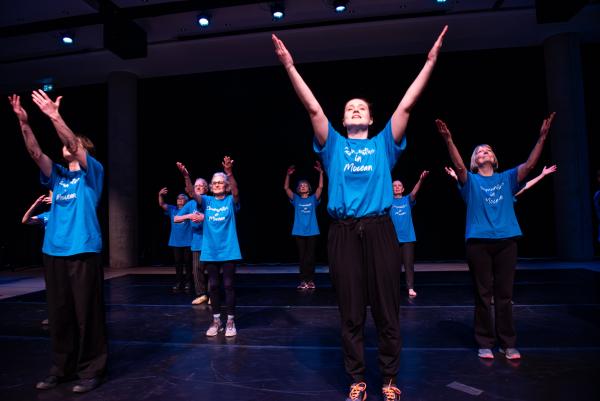 Elderly dancers in blue shirts against black background, arms reaching