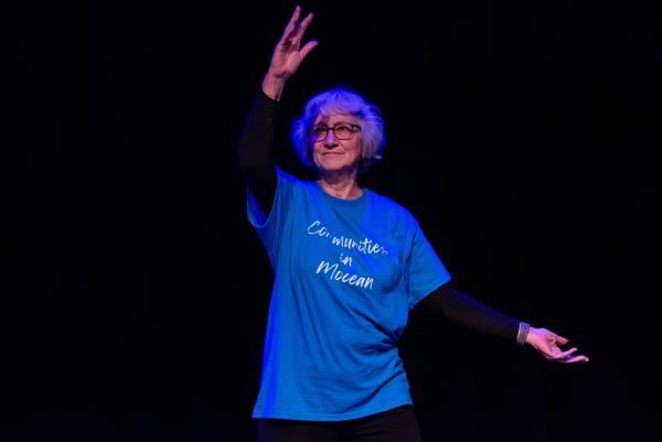 Elderly dancer in blue shirt against black background, arms reaching