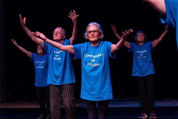 a group of older adults in blue t-shirts stand on a stage with arms outstretched in a V