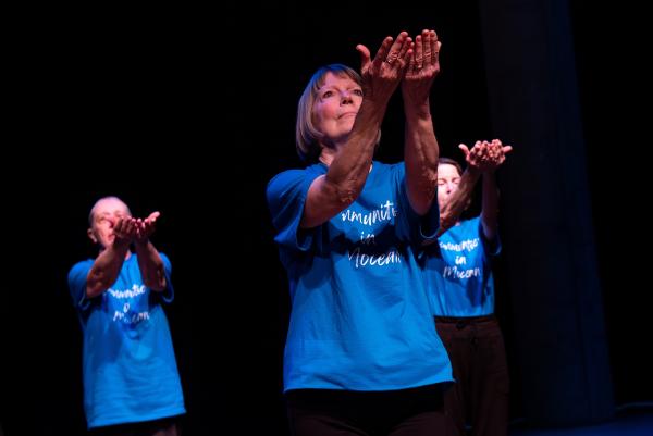 Elderly dancers in blue shirts against black background, arms reaching
