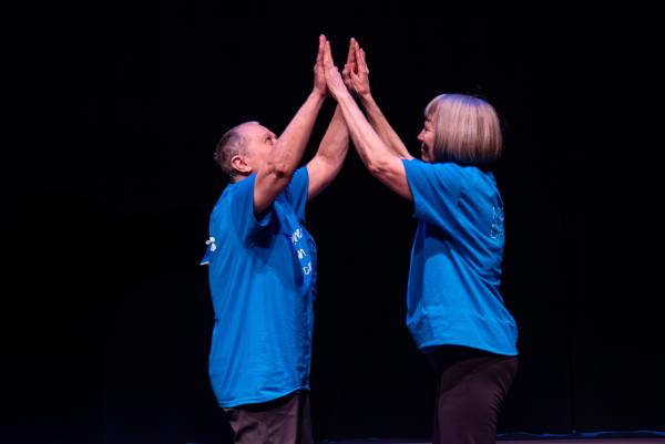 Elderly dancer in blue shirt against black background, arms reaching