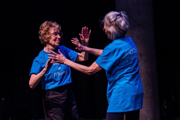 Elderly dancers in blue shirts against black background, arms reaching